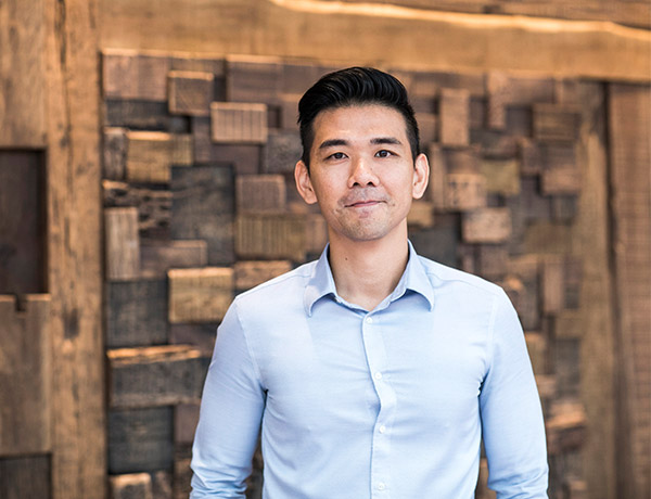 Standing man in blue shirt with wooden wall backdrop.