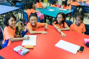 Smiling children wearing orange shirts sitting at a table with notebooks