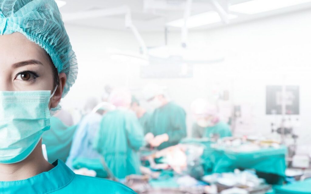 A woman in a scrub suit stands confidently in front of a hospital, ready to provide medical care.