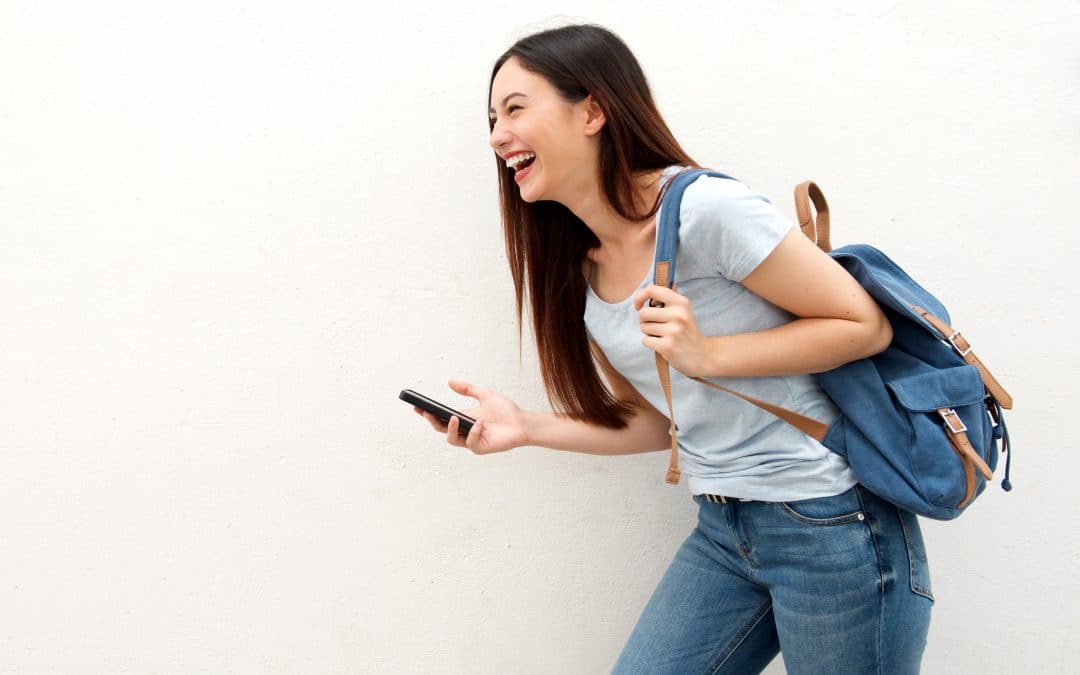 A woman joyfully laughs while carrying a backpack and holding a phone in her hand.