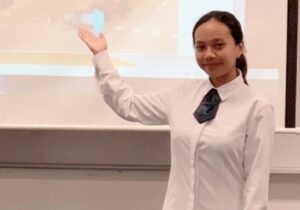 A professional woman in a white shirt and tie stands confidently in front of a projector, ready to deliver a presentation.